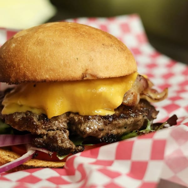 A juicy burger with melted cheese, bacon, red onion, and lettuce on a bun, in a red and white checkered paper.