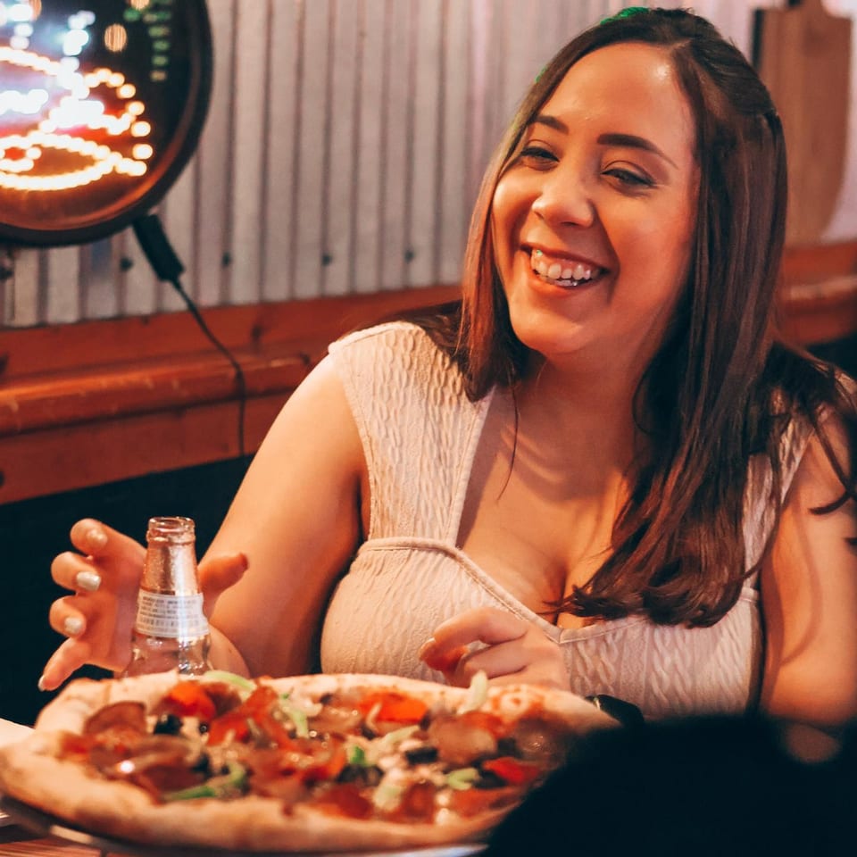 A smiling woman enjoying a pepperoni and vegetable pizza with a drink at Up North Pizzeria.