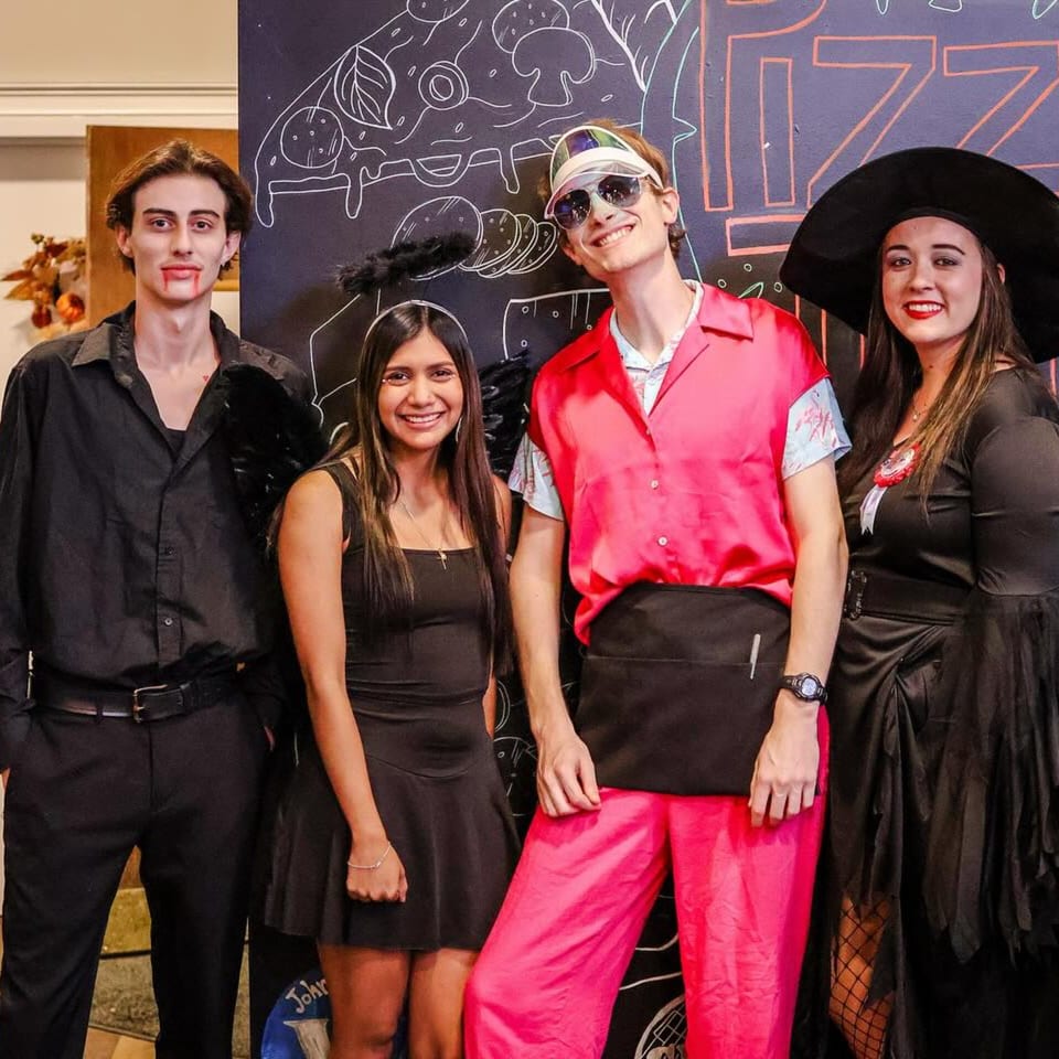 Four people in Halloween costumes, including a vampire, witch, and a person in a pink outfit, smiling.