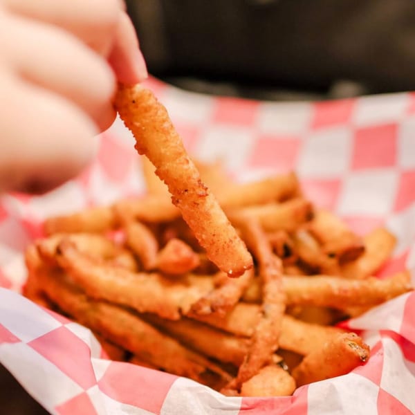 A hand picks up a crispy french fry from a red and white checkered paper basket.