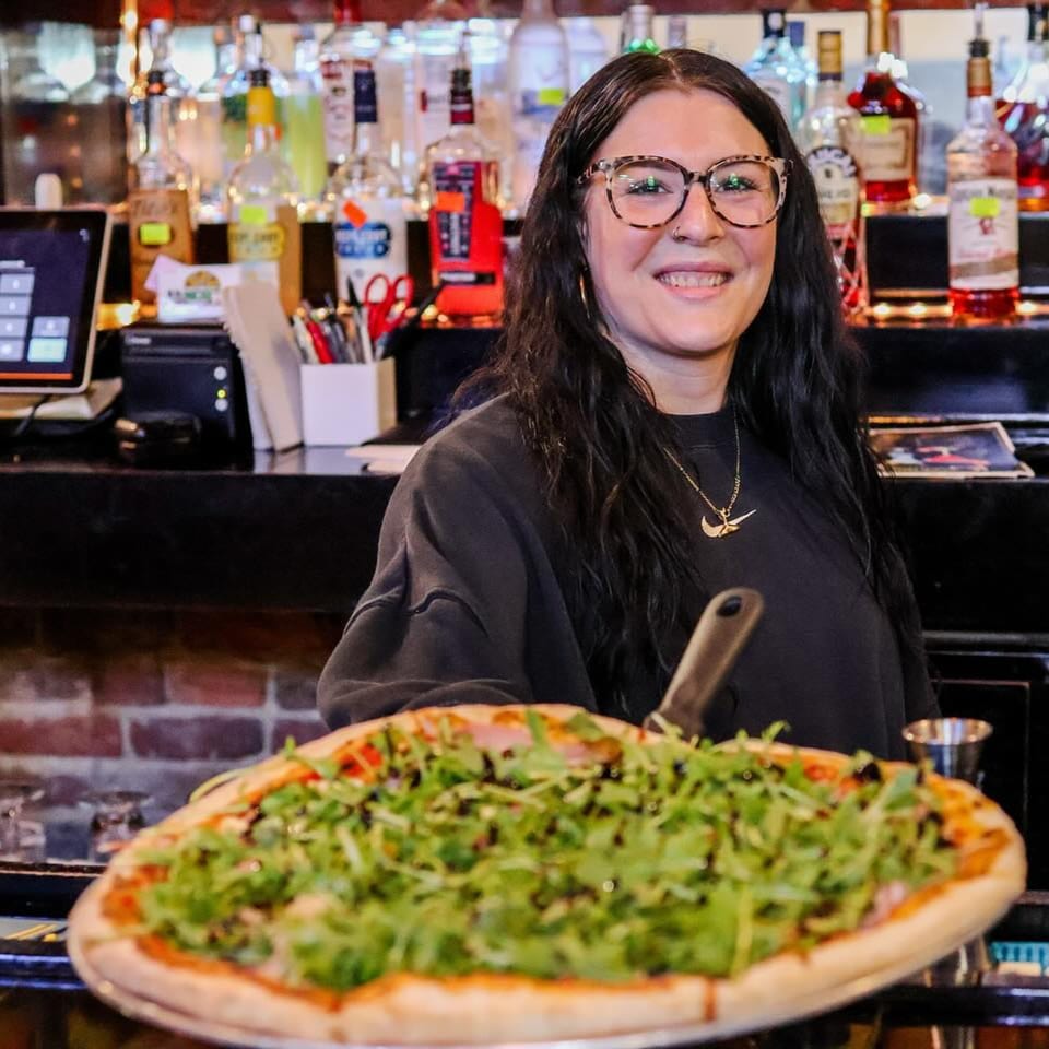 A smiling Up North Pizzeria employee holding a freshly made pizza topped with arugula and balsamic glaze.
