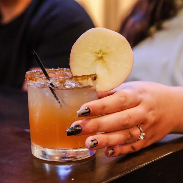 A hand with painted nails holds an apple cider cocktail garnished with an apple slice and cinnamon sugar rim.