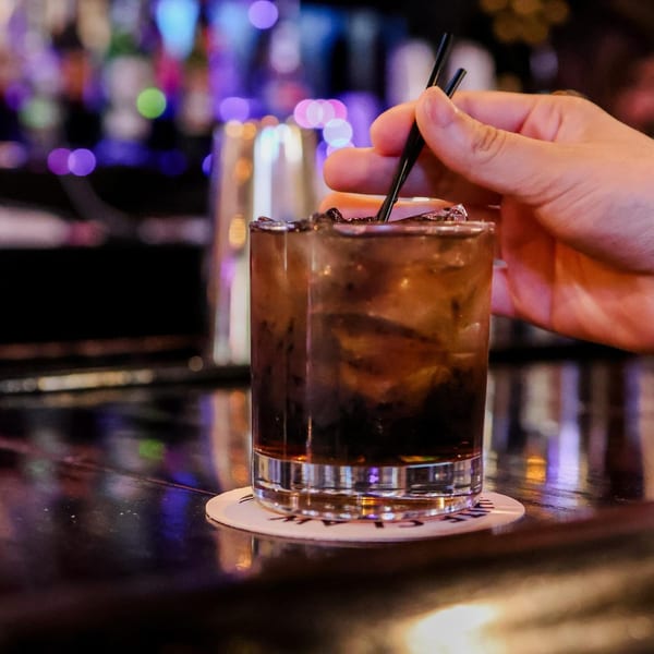 Hand stirring a dark cocktail with ice and two black straws on a bar with blurred lights.