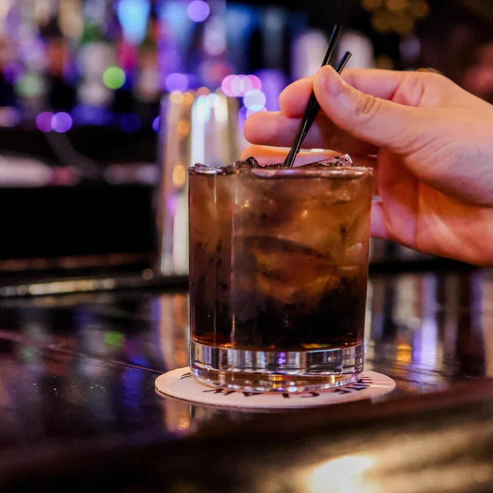 Hand stirring a dark cocktail with ice and two black straws on a bar with blurred lights.