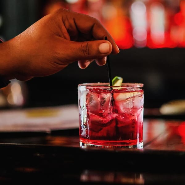 A hand stirring a vibrant red cocktail with ice and lime in a glass on a dark bar.