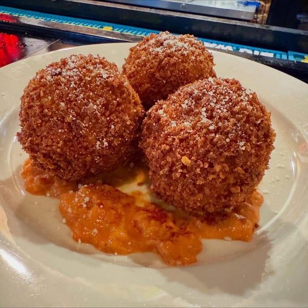 Three golden-brown arancini balls, sprinkled with cheese, resting on a bed of orange sauce.