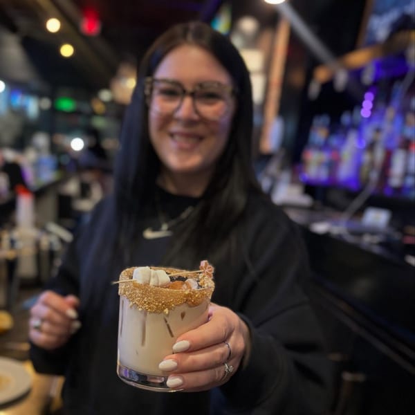 Smiling woman in glasses holding a s'mores cocktail with a graham cracker rim and marshmallows.