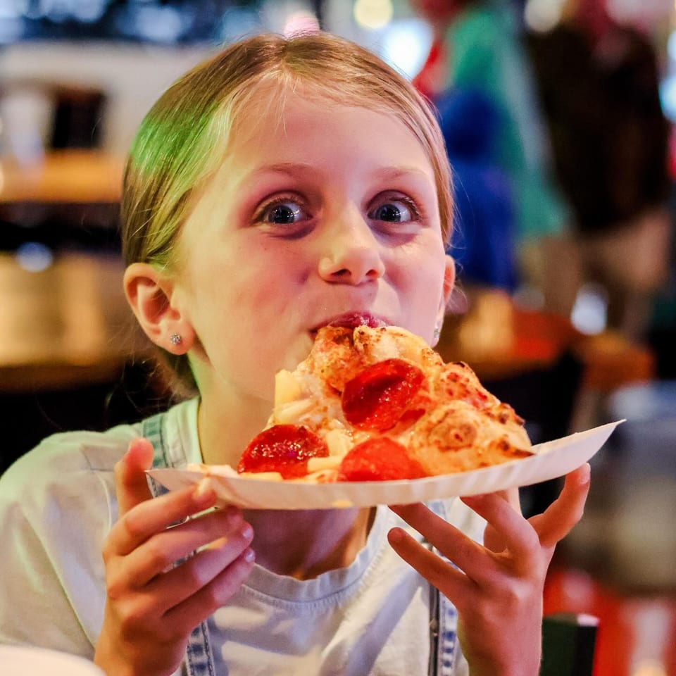 A young girl with blonde hair takes a big bite out of a slice of pepperoni pizza.