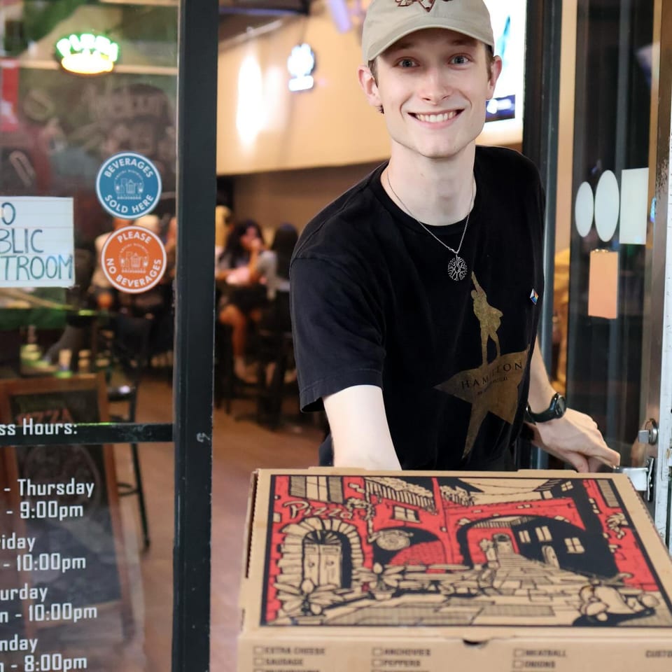 Smiling Up North Pizzeria employee in a cap and black shirt holding a pizza box.